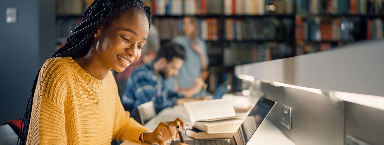 Universitätsbibliothek: Begabtes schwarzes Mädchen benutzt Laptop und macht Notizen