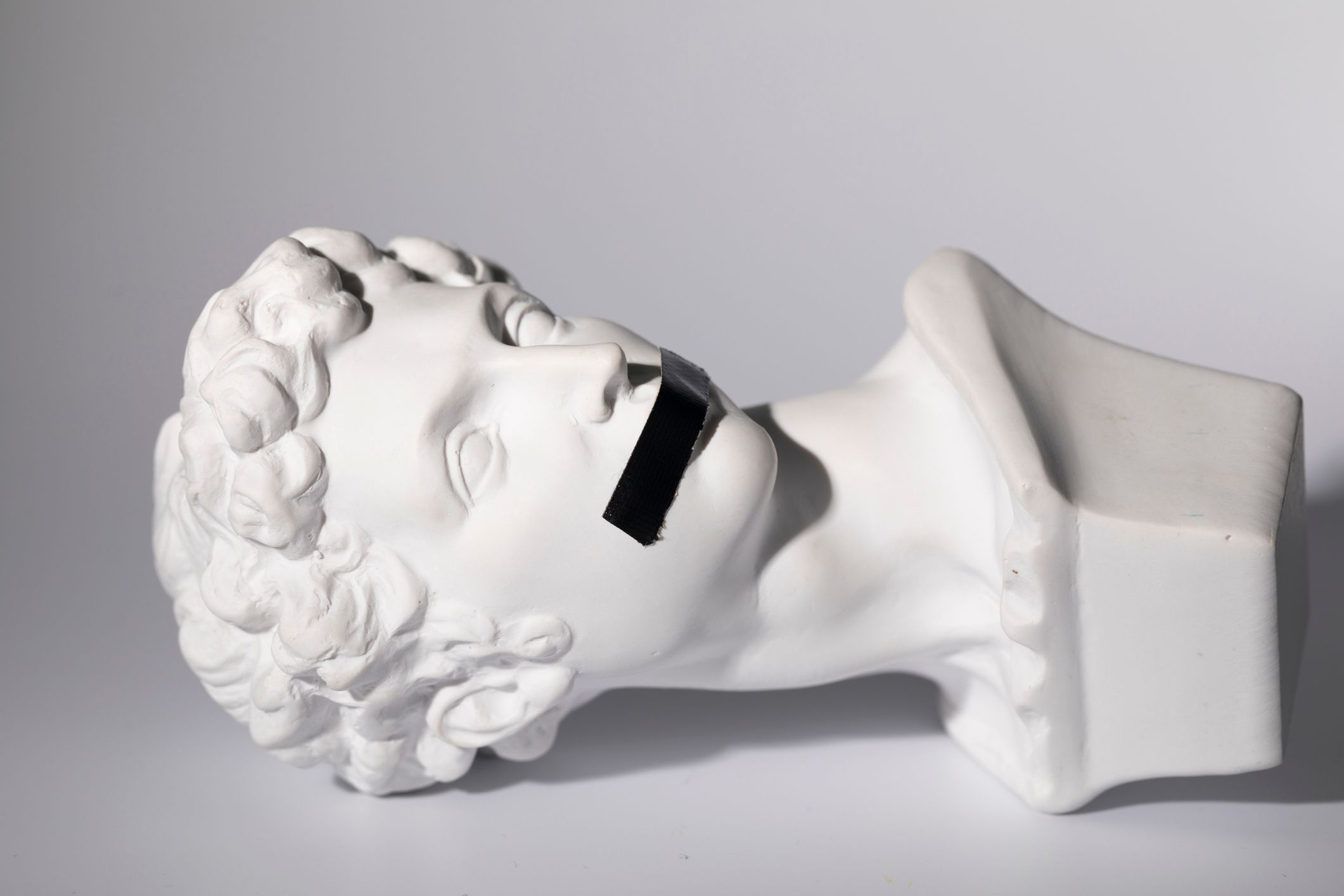 Foto einer mittelalterlichen weißen Steinskulptur des Kopfes eines jungen Mannes, dessen Mund mit schwarzem Klebeband bedeckt ist.