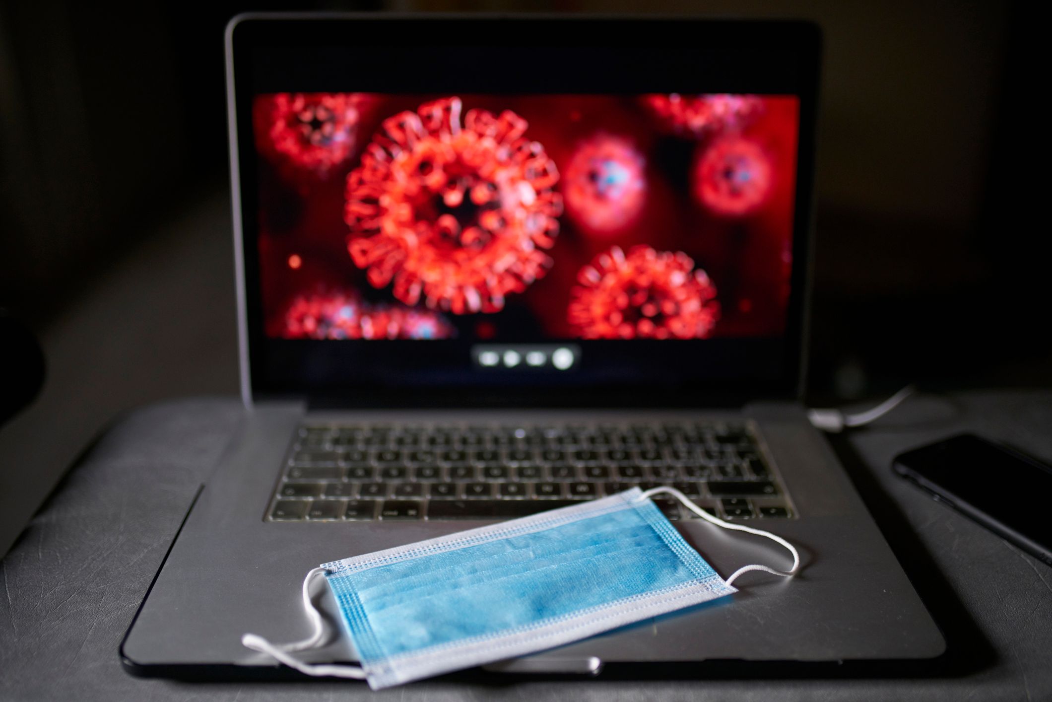 In einem dunklen Raum liegt eine blaue Gesichtsmaske auf einer Laptop-Tastatur, während auf dem Bildschirm mikroskopische Aufnahmen des stacheligen roten COVID-Virus zu sehen sind.