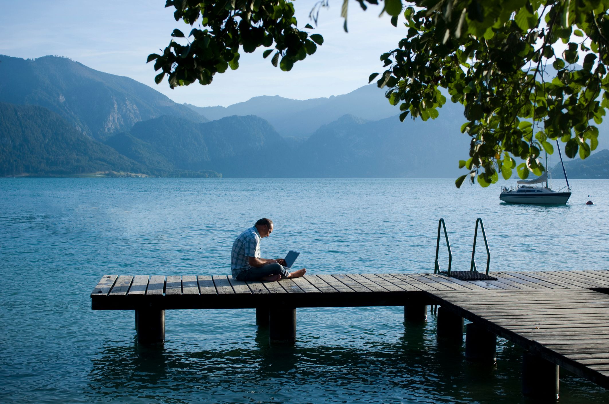 Foto eines Mannes, der mit gekreuzten Beinen auf einem Steg an einem See sitzt und einen Laptop auf dem Schoß hat.