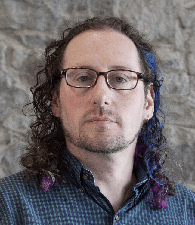 Porträtfoto des Autors. Ein weißer Mann im Alter von 30s oder 40s Jahren, der eine Brille trägt und in dessen lockigem braunem Haar ein Hauch von Blau zu erkennen ist.