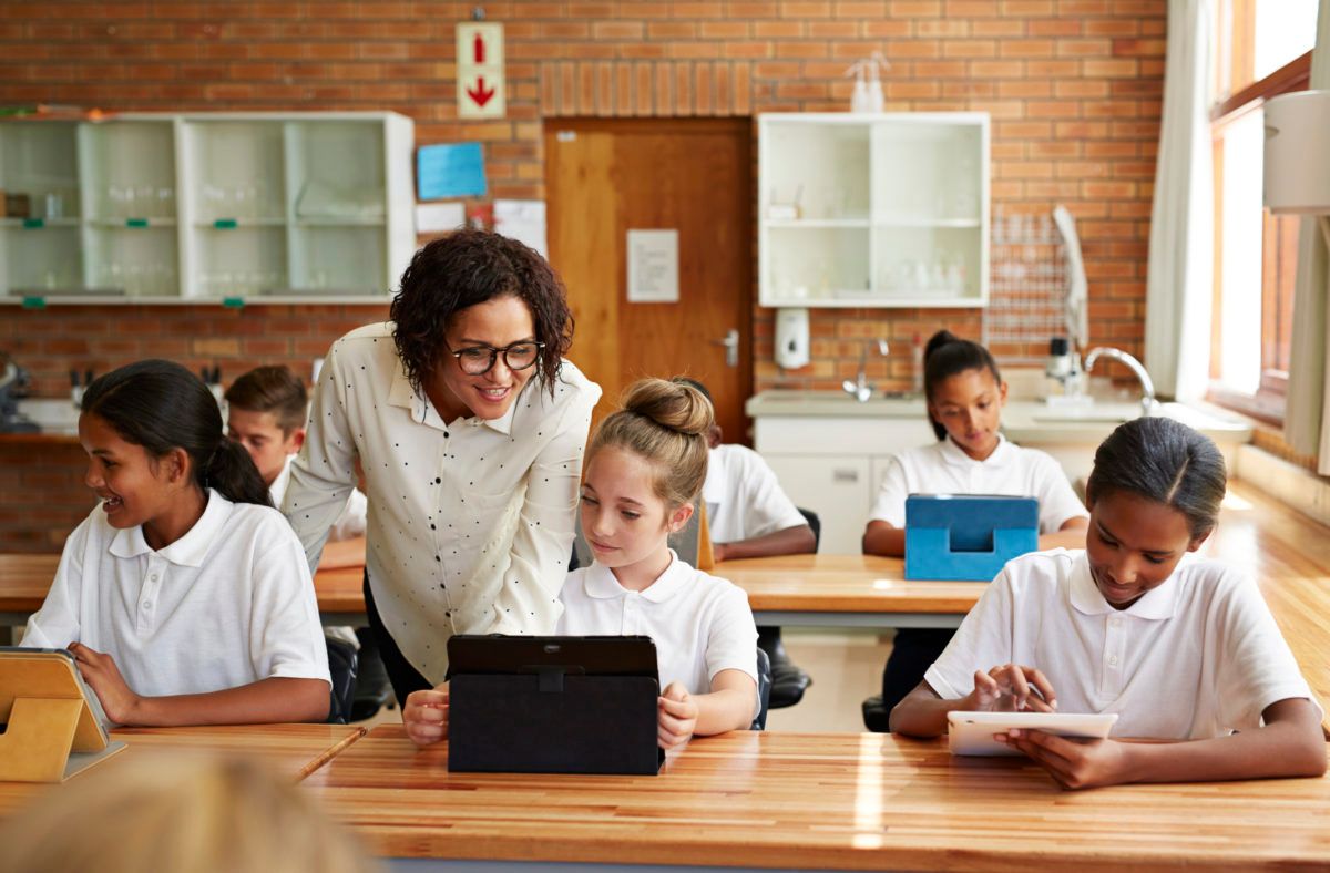 Lehrer, der Schülern an ihren Schreibtischen im Klassenzimmer mit einem Tablet und Unterlagen hilft
